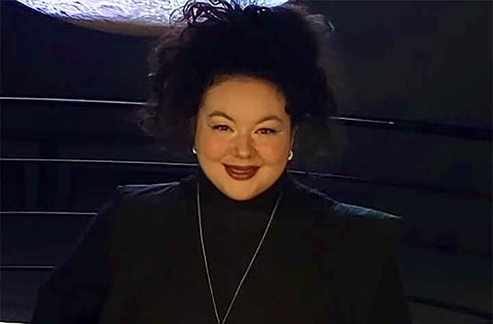Smiling woman with curly dark hair wearing a black turtleneck and blazer, standing indoors against a dark background. She is wearing earrings and a long necklace, looking directly at the camera.