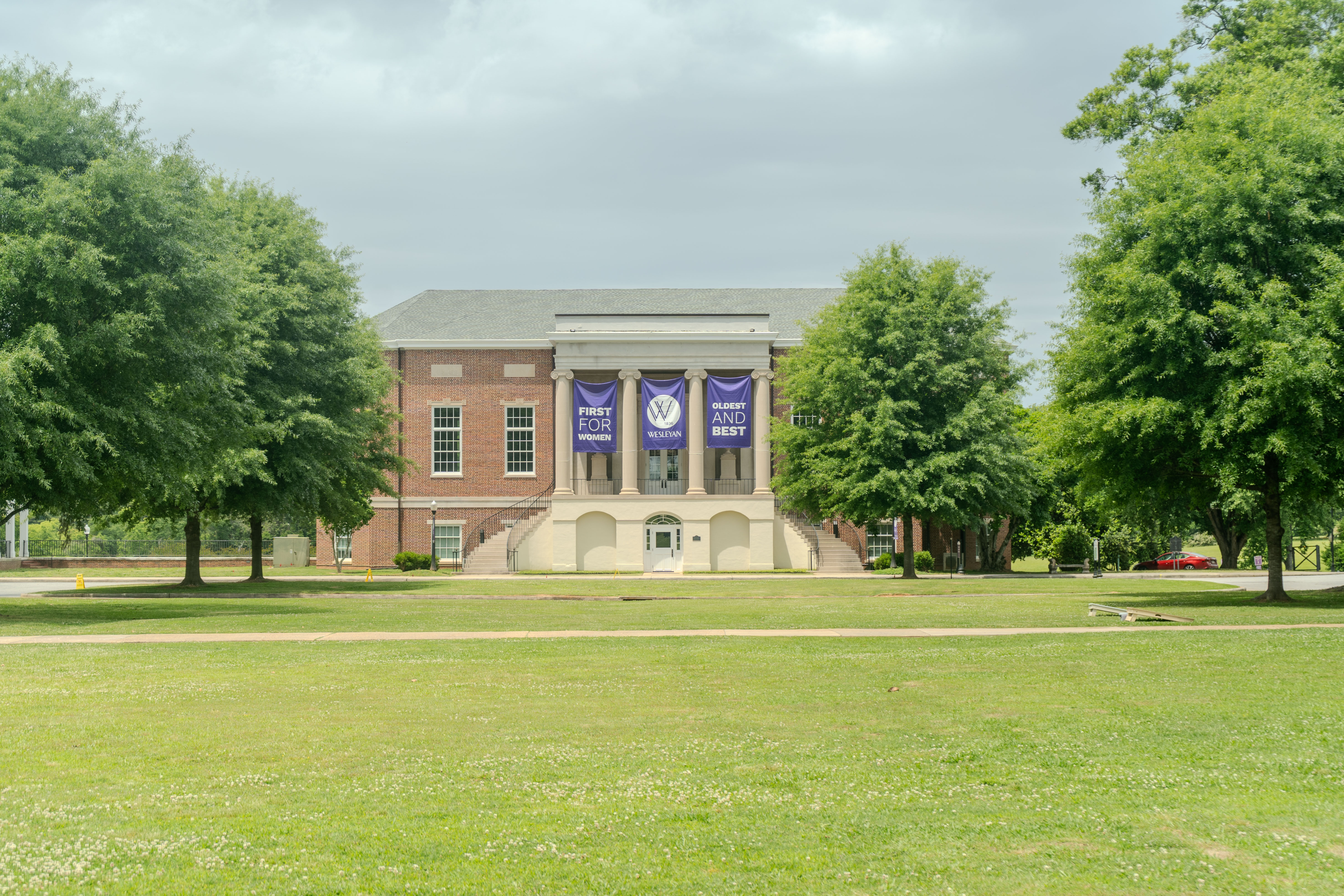 Front view of a stately brick campus building with white columns, centered across a green lawn and framed by large trees. Purple banners hang between the columns, including one with the Wesleyan logo and others reading “First for Women” and “Oldest and Be