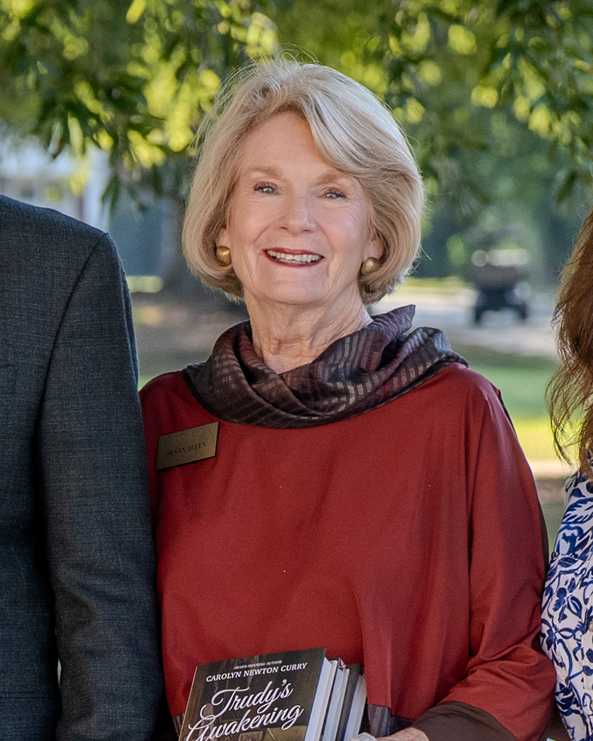 Smiling older woman with short blonde hair wearing a red blouse and patterned scarf, standing outdoors under leafy trees. She has gold earrings and a name badge that reads “Susan Allen.” She is holding several copies of a book titled Trudy’s Awakening by 
